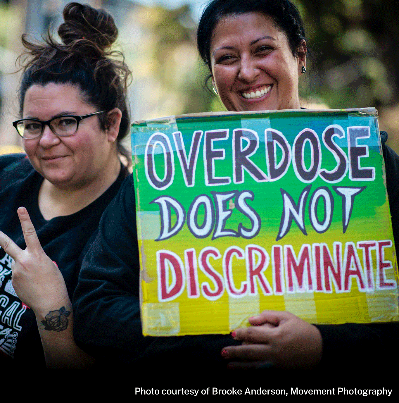 Two people pose together. Person at left smiles while making peace sign hand gesture. They have black shirt on that reads in part, “RADICAL LOVE” with red flowers. They have a tattoo of a flower near their left wrist. Person at right wears black sweatshirt and smiles while holding sign reading, OVERDOSE DOES NOT DISCRIMINATE.